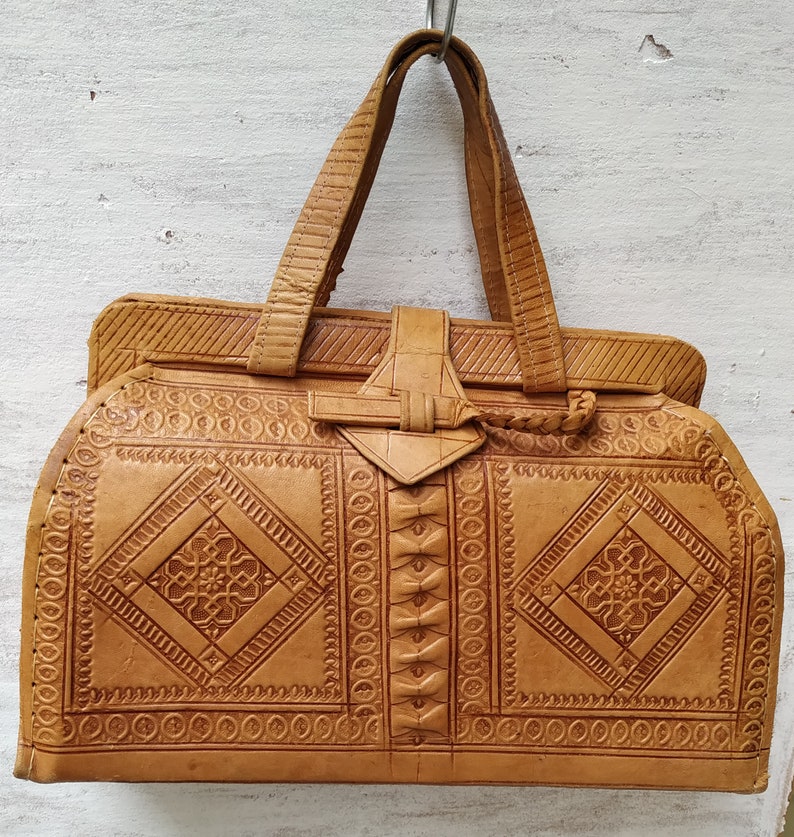 Puede incluir: Un bolso de mano de cuero marr&oacute;n con intrincados patrones en relieve. El bolso tiene un asa superior y un cierre de solapa con una hebilla decorativa.