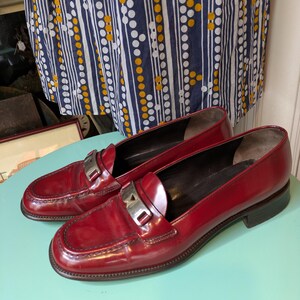 May include: A pair of red leather loafers with silver-tone metal accents. The shoes have a classic design with a rounded toe and a low heel. The loafers are displayed on a light blue surface.