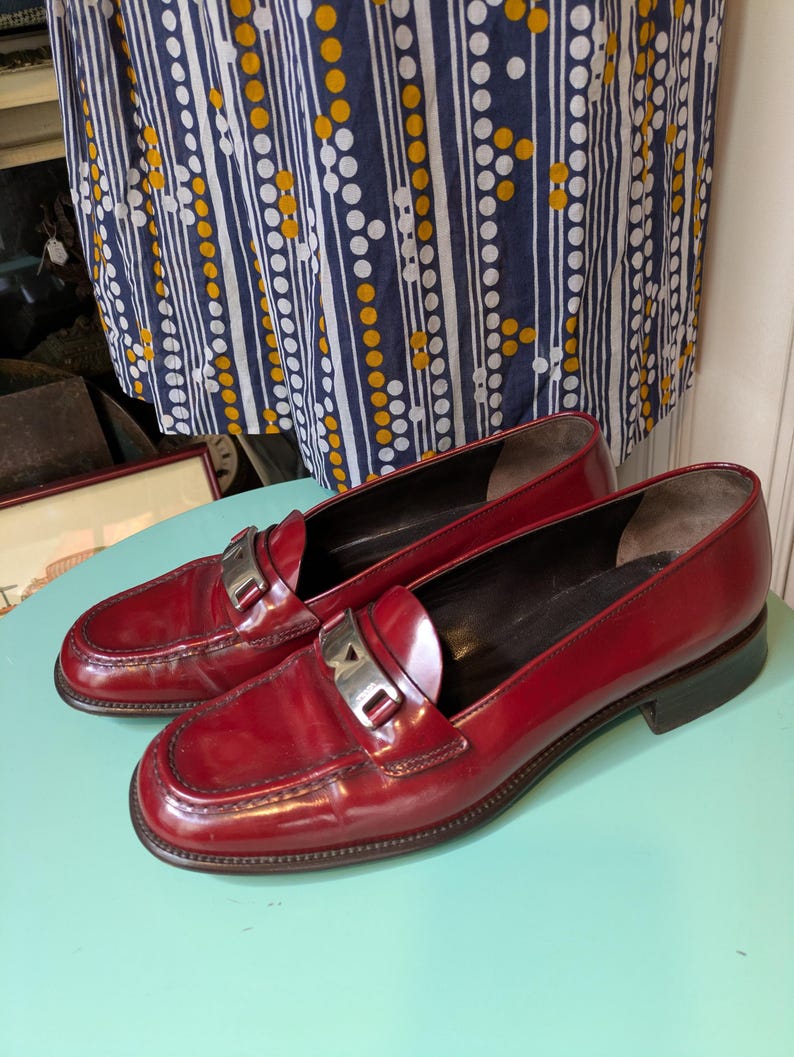 May include: A pair of red leather loafers with silver-tone metal accents. The shoes have a classic design with a rounded toe and a low heel. The loafers are displayed on a light blue surface.