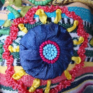 May include: A close-up of a colorful embroidered fabric with a blue flower in the center. The flower is surrounded by yellow, red, and green fabric. The fabric is textured and has a striped pattern.