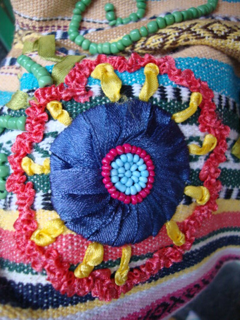 May include: A close-up of a colorful embroidered fabric with a blue flower in the center. The flower is surrounded by yellow, red, and green fabric. The fabric is textured and has a striped pattern.