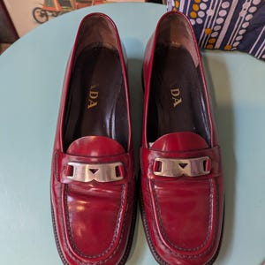 May include: A pair of red leather loafers with silver-tone hardware. The shoes have a classic design with a rounded toe and a low heel. The word "ADA" is visible on the inner sole of each shoe. The loafers are displayed on a light blue surface.