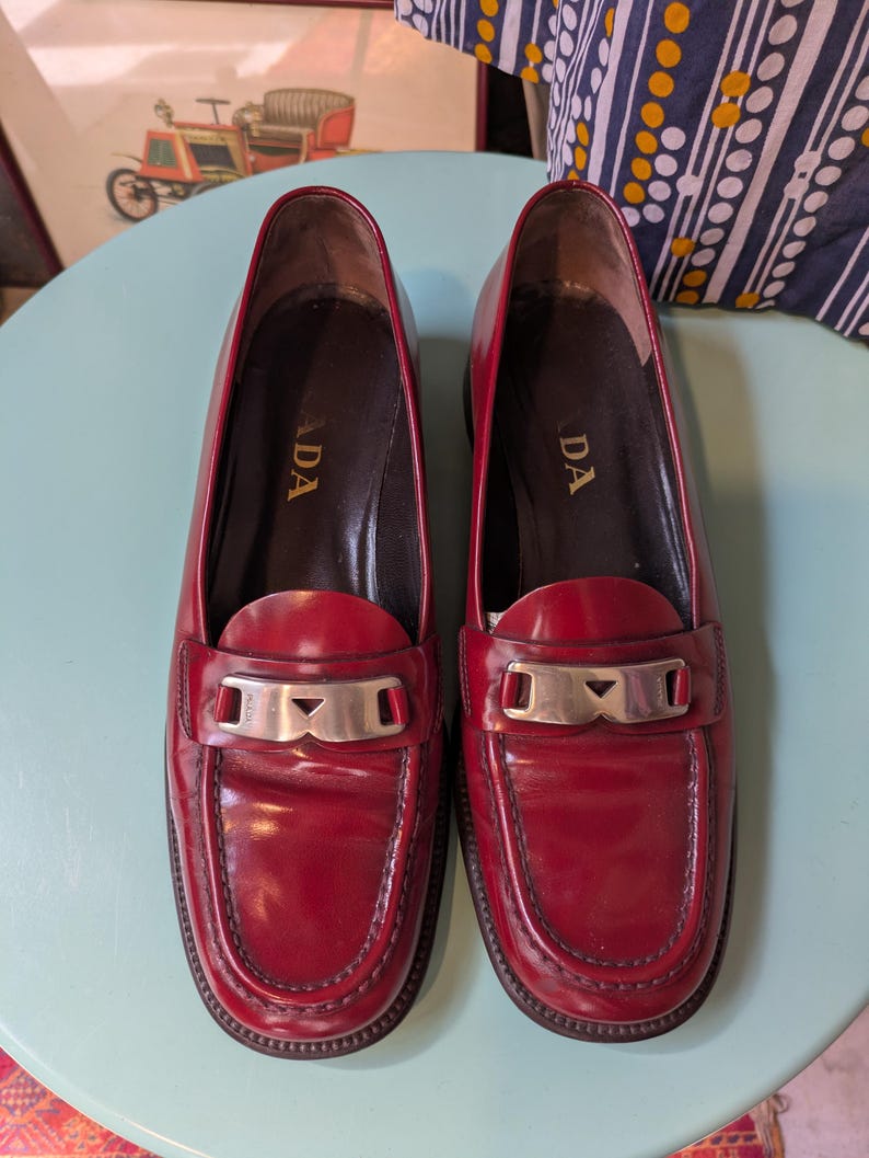 May include: A pair of red leather loafers with silver-tone hardware. The shoes have a classic design with a rounded toe and a low heel. The word "ADA" is visible on the inner sole of each shoe. The loafers are displayed on a light blue surface.
