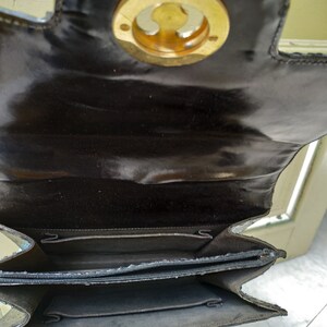 Puede incluir: Un bolso de mano de cuero negro con un cierre circular dorado. El bolso est&aacute; abierto y el interior est&aacute; forrado con una tela oscura. El bolso tiene una cremallera.