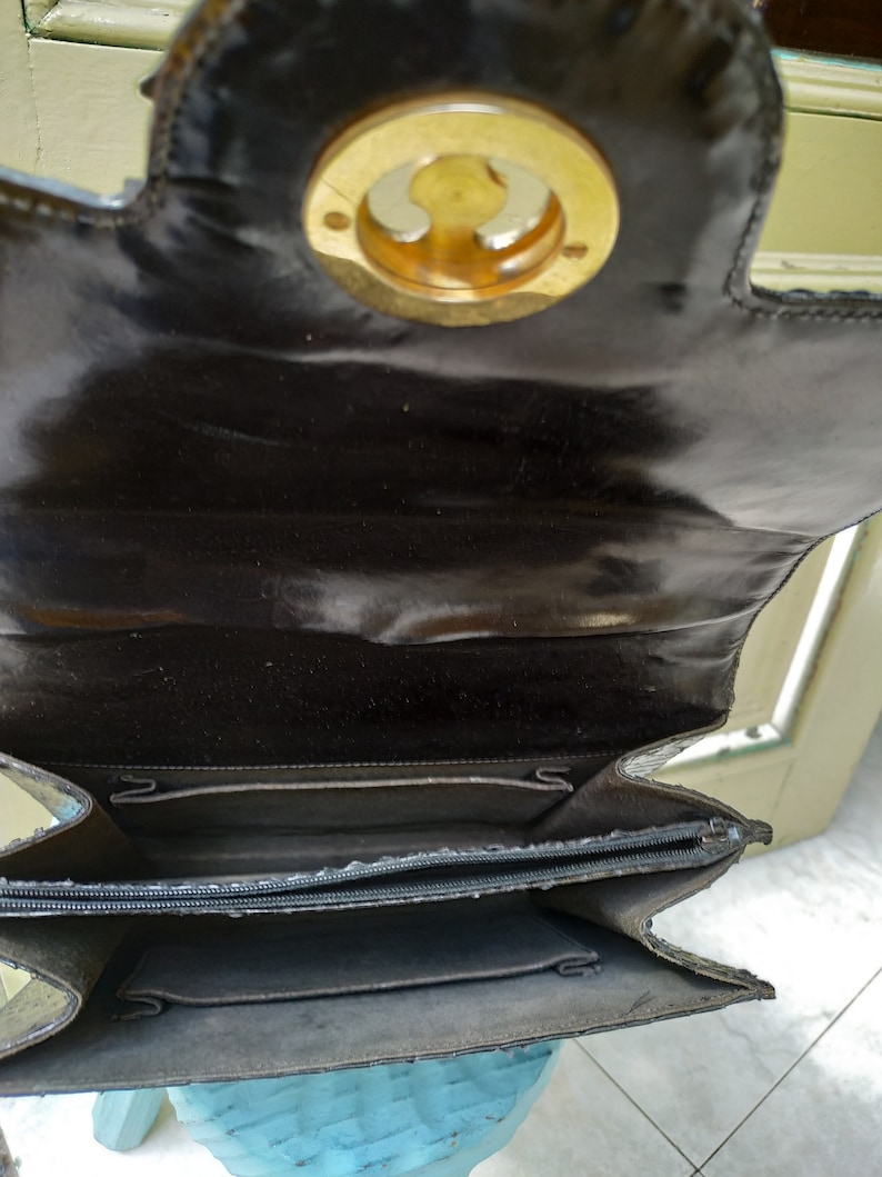 Puede incluir: Un bolso de mano de cuero negro con un cierre circular dorado. El bolso est&aacute; abierto y el interior est&aacute; forrado con una tela oscura. El bolso tiene una cremallera.