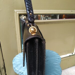 Puede incluir: Un bolso de mano de piel de serpiente negra con cierre dorado y asa superior. El bolso est&aacute; sobre un taburete azul.