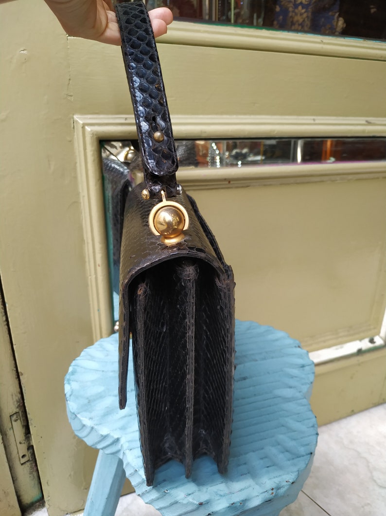 Puede incluir: Un bolso de mano de piel de serpiente negra con cierre dorado y asa superior. El bolso est&aacute; sobre un taburete azul.
