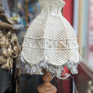 Op de afbeelding: Een witte gehaakte lampenkap met een franje zit op een houten voet. De lampenkap heeft een delicaat, ingewikkeld patroon.