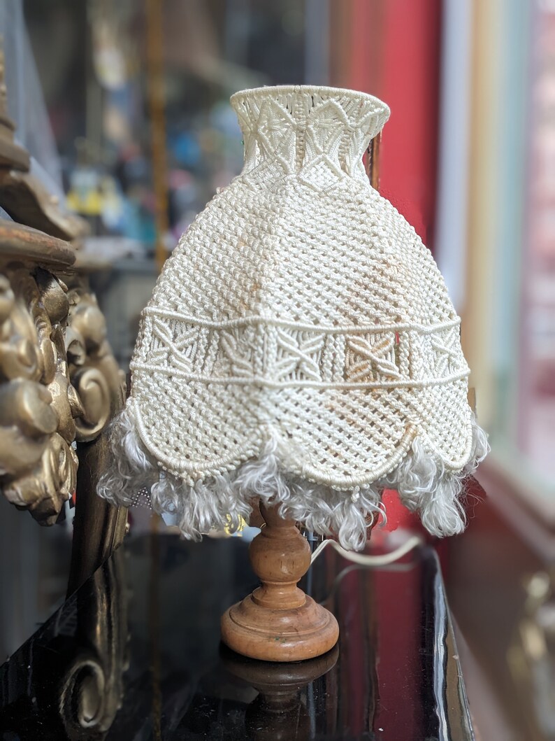 Op de afbeelding: Een witte gehaakte lampenkap met een franje zit op een houten voet. De lampenkap heeft een delicaat, ingewikkeld patroon.
