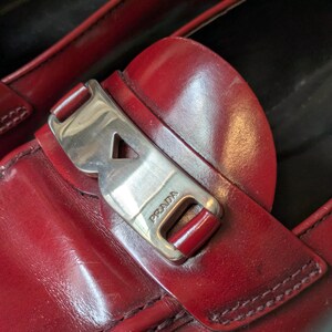 May include: Close-up of a pair of red leather loafers. The shoes feature a silver-tone buckle with the brand name "PRADA" and visible stitching. The leather has a glossy finish.