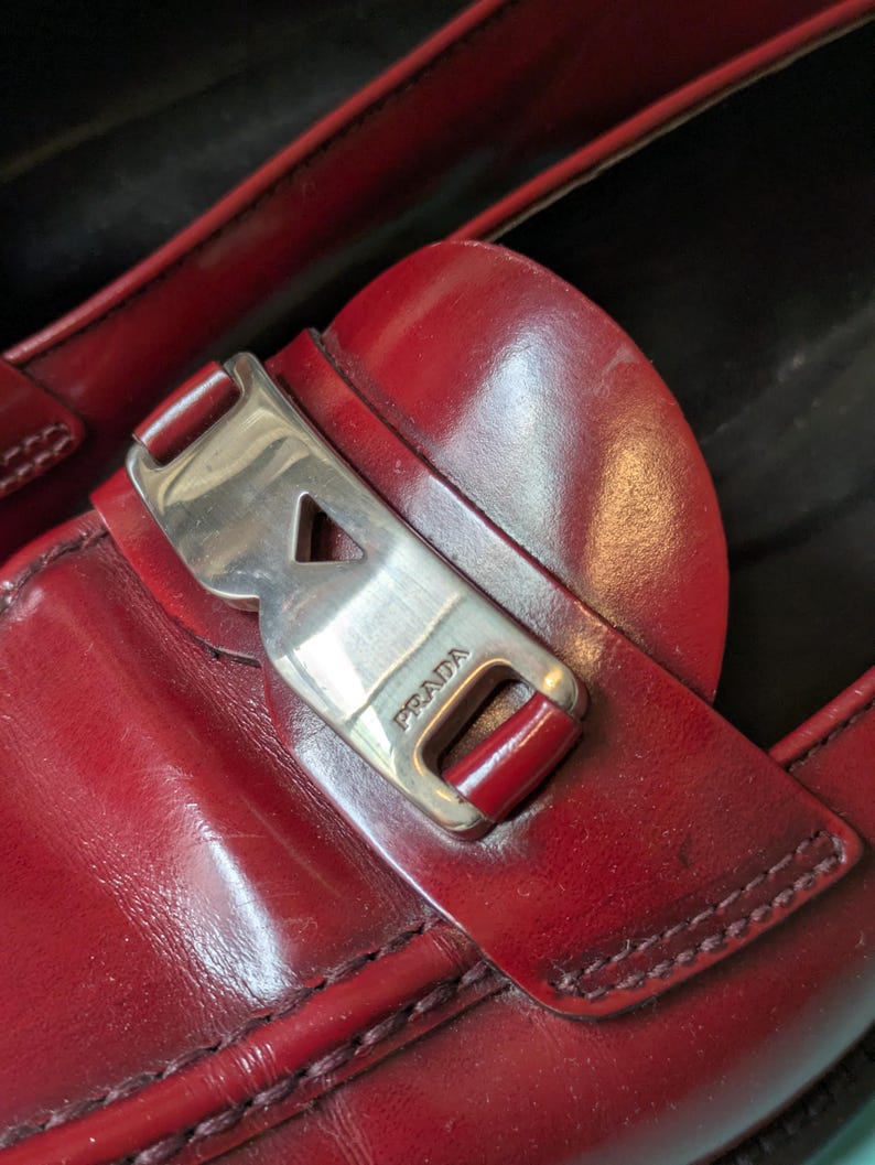 May include: Close-up of a pair of red leather loafers. The shoes feature a silver-tone buckle with the brand name "PRADA" and visible stitching. The leather has a glossy finish.