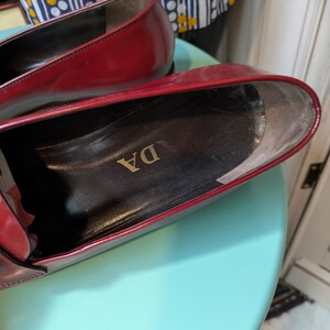 May include: Close-up of a pair of red leather loafers. The interior of the shoe is black with gold lettering. The shoes are on a light blue surface.