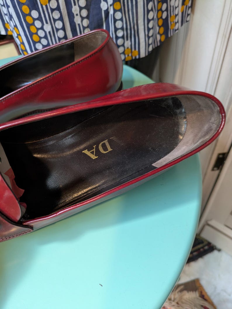 May include: Close-up of a pair of red leather loafers. The interior of the shoe is black with gold lettering. The shoes are on a light blue surface.