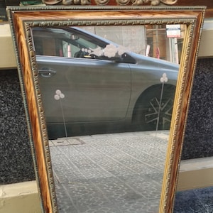 May include: A vintage-style mirror with a gold-toned frame and a wood-grain finish. The frame has a decorative design at the top and the mirror reflects a car and a tiled floor.