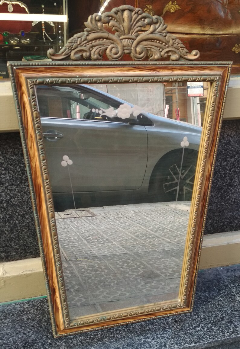 May include: A vintage-style mirror with a gold-toned frame and a wood-grain finish. The frame has a decorative design at the top and the mirror reflects a car and a tiled floor.