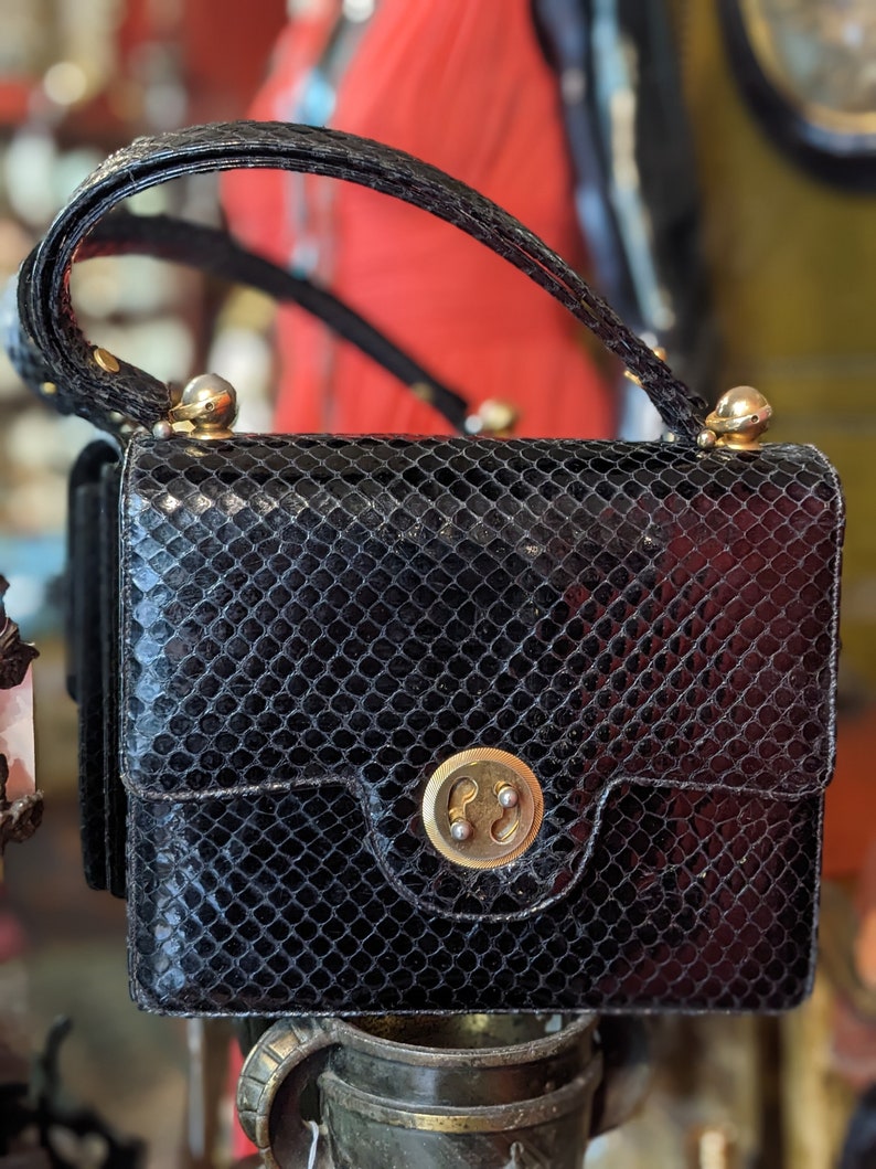 Puede incluir: Un bolso de mano de piel de serpiente negra con cierre dorado y asa superior. El bolso tiene una superficie texturizada y escamosa.