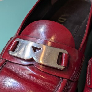 May include: Close-up of a red leather loafer with a silver-tone buckle. The shoe features a rounded toe and a stitched design. The buckle is rectangular with the brand name engraved. The leather has a glossy finish.