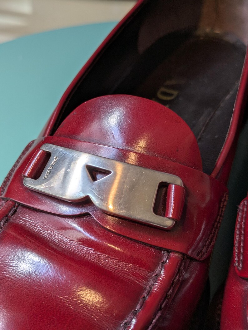 May include: Close-up of a red leather loafer with a silver-tone buckle. The shoe features a rounded toe and a stitched design. The buckle is rectangular with the brand name engraved. The leather has a glossy finish.