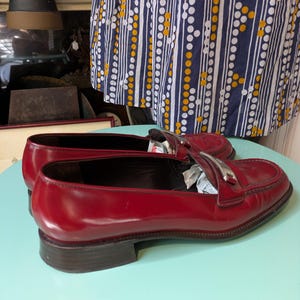 May include: A pair of glossy red leather loafers with silver-tone accents. The shoes have a low heel and a classic design. The loafers are displayed on a light blue surface.