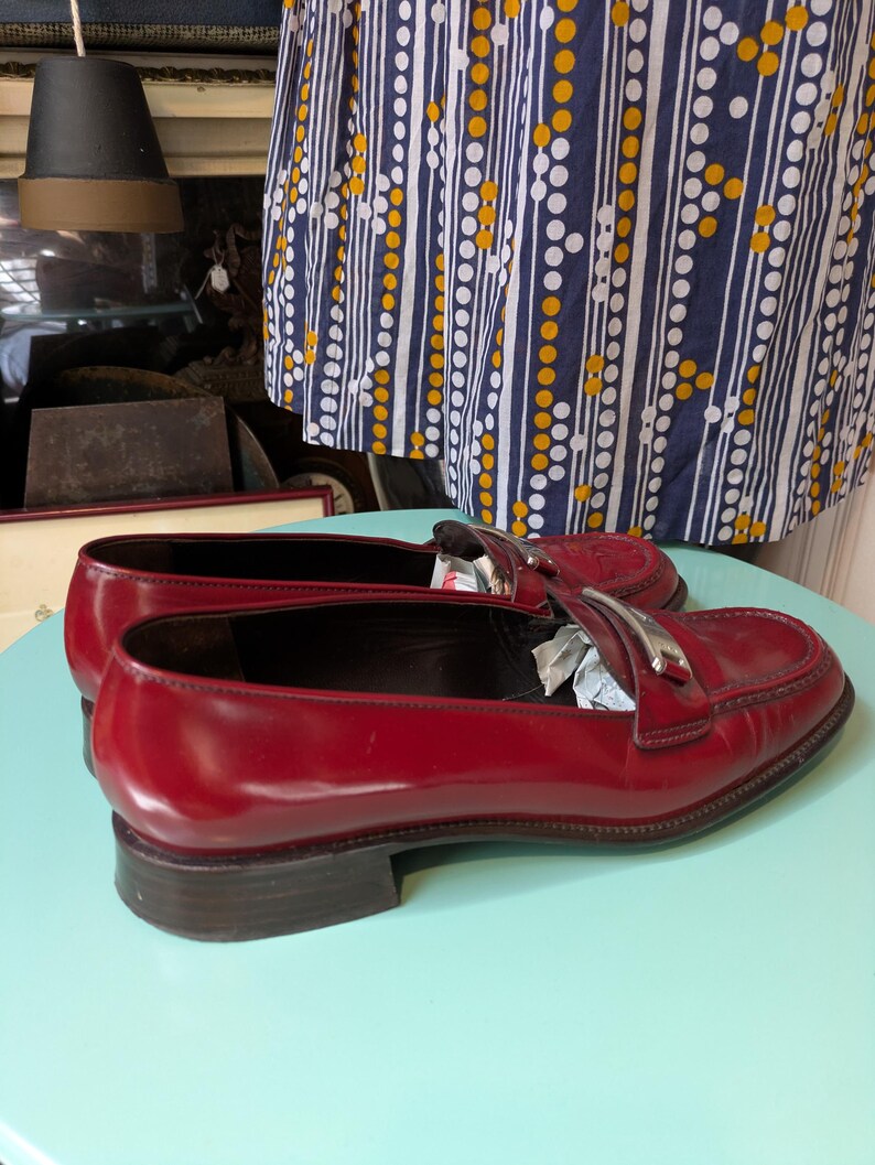 May include: A pair of glossy red leather loafers with silver-tone accents. The shoes have a low heel and a classic design. The loafers are displayed on a light blue surface.