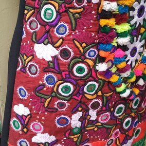 May include: A red, orange, green, and yellow embroidered fabric with colorful pom-poms and small round mirrors.