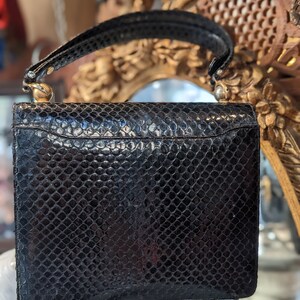 Puede incluir: Un bolso de mano de piel de serpiente negra con cierre y asa dorados. El bolso tiene una superficie texturizada y una forma rectangular.