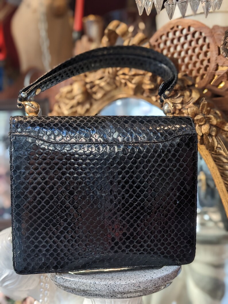 Puede incluir: Un bolso de mano de piel de serpiente negra con cierre y asa dorados. El bolso tiene una superficie texturizada y una forma rectangular.