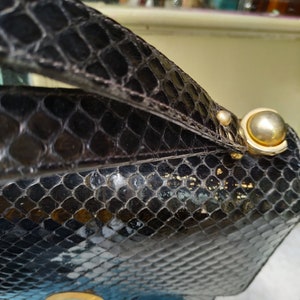 Puede incluir: Un bolso de mano de piel de serpiente negra con cierre dorado y asa dorada. El bolso tiene una superficie texturizada y un acabado brillante.