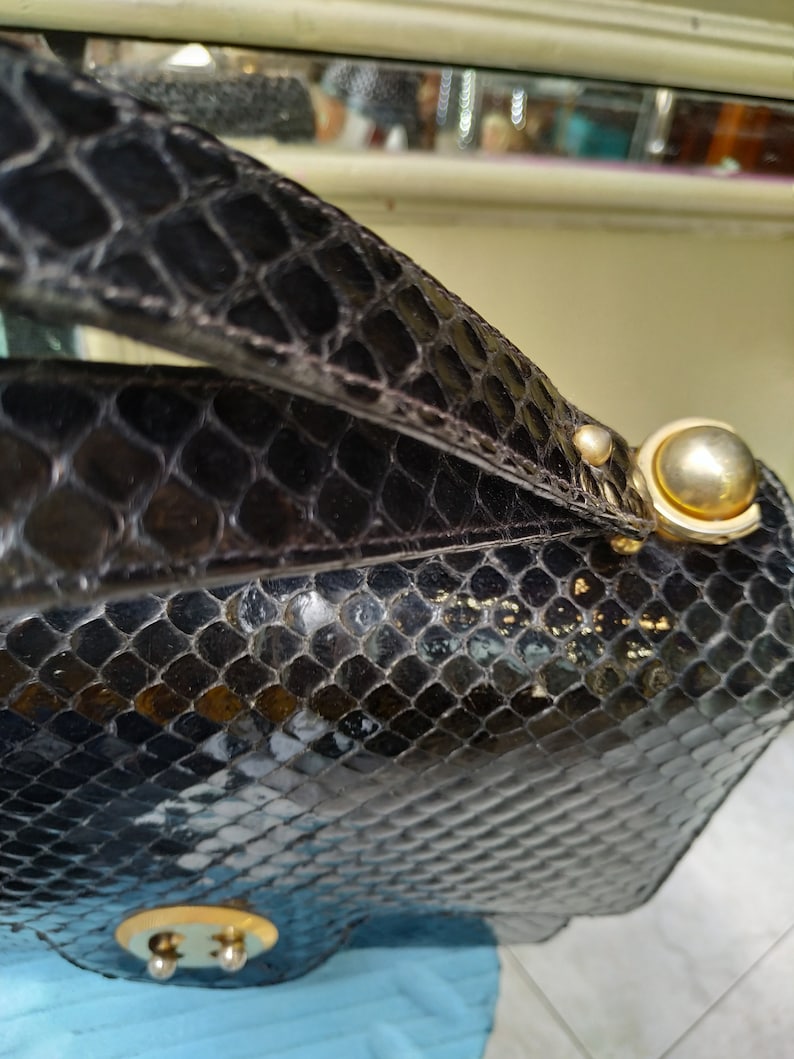 Puede incluir: Un bolso de mano de piel de serpiente negra con cierre dorado y asa dorada. El bolso tiene una superficie texturizada y un acabado brillante.