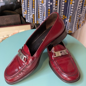 May include: A pair of glossy, burgundy leather loafers with silver-tone metal accents. The shoes have a classic design with a low heel and a decorative strap across the vamp. The shoes are displayed on a light blue surface.