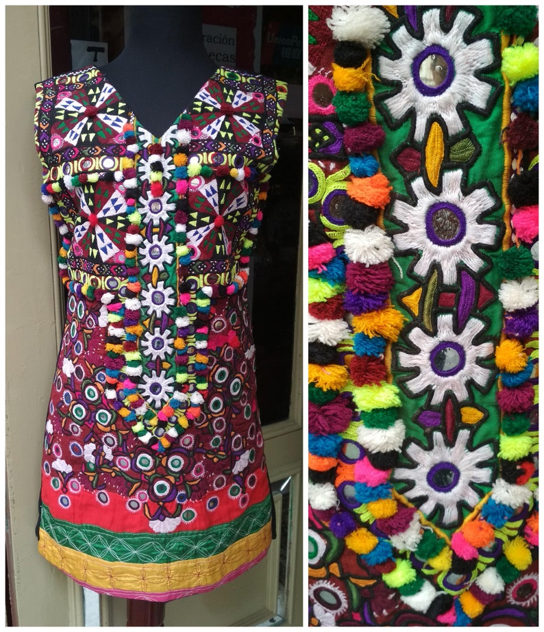 May include: A colorful sleeveless tunic with intricate embroidery, mirrors, and pom-poms. The tunic is made of a red, green, and black fabric with a geometric pattern. The embroidery is in a variety of colors, including white, blue, yellow, and pink.