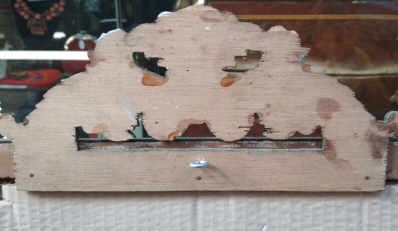 May include: The back of a wooden mirror frame with a decorative, ornate design. The frame is made of light wood with a natural finish. There is a small metal hook on the back of the frame.