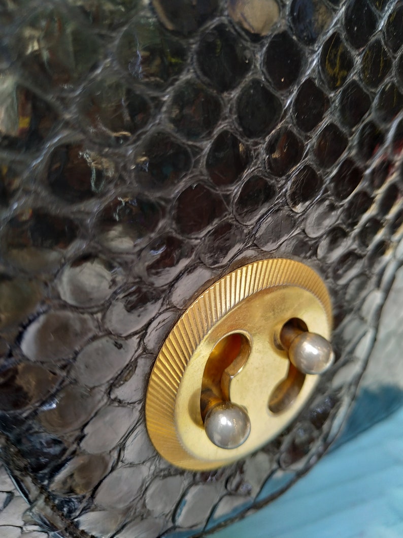 Puede incluir: Primer plano de un bolso de piel de serpiente negro con un cierre dorado. El cierre tiene dos peque&ntilde;os remaches redondos de color plateado.