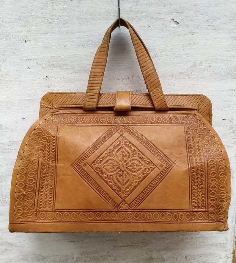 Puede incluir: Un bolso de mano de cuero marr&oacute;n con intrincados dise&ntilde;os en relieve. El bolso tiene un asa superior y un cierre de solapa.