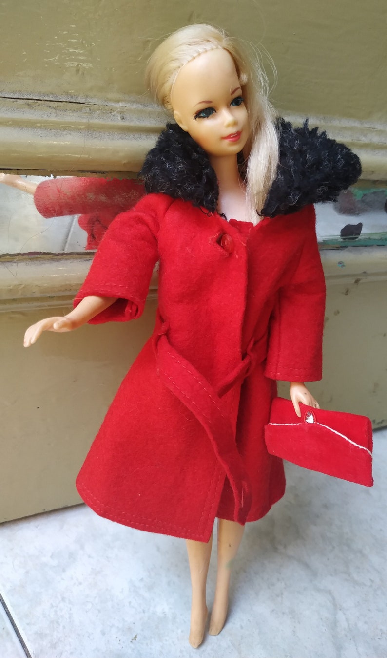 May include: A red coat with a black fur collar and a red handbag. The coat has a belt and a button fastening. The doll is wearing the coat and holding the handbag.