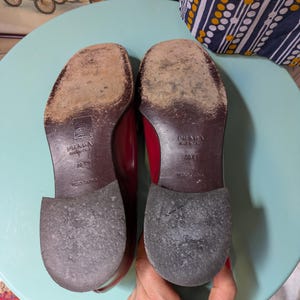 May include: A pair of red Prada leather shoes, soles facing up, showing wear and the brand's logo. The shoes are size 38 1/2, made in Italy. The background includes a vintage car painting and a blue and white patterned fabric.