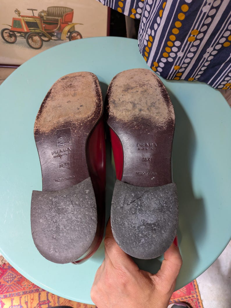 May include: A pair of red Prada leather shoes, soles facing up, showing wear and the brand's logo. The shoes are size 38 1/2, made in Italy. The background includes a vintage car painting and a blue and white patterned fabric.