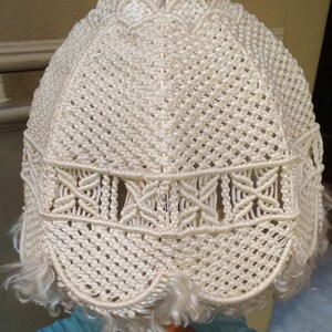 Op de afbeelding: Een witte macram&eacute; lampenkap met een houten voet en een wit snoer. De lampenkap heeft een franje rond de onderkant.