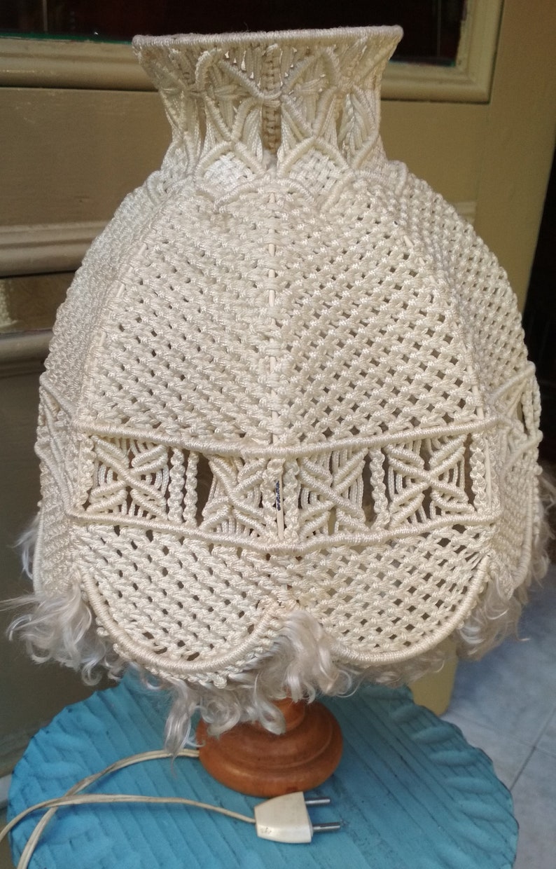 Op de afbeelding: Een witte macram&eacute; lampenkap met een houten voet en een wit snoer. De lampenkap heeft een franje rond de onderkant.
