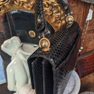 Puede incluir: Un bolso de mano de piel de serpiente negra con asa superior y herrajes dorados. El bolso tiene forma rectangular y cierre de solapa.