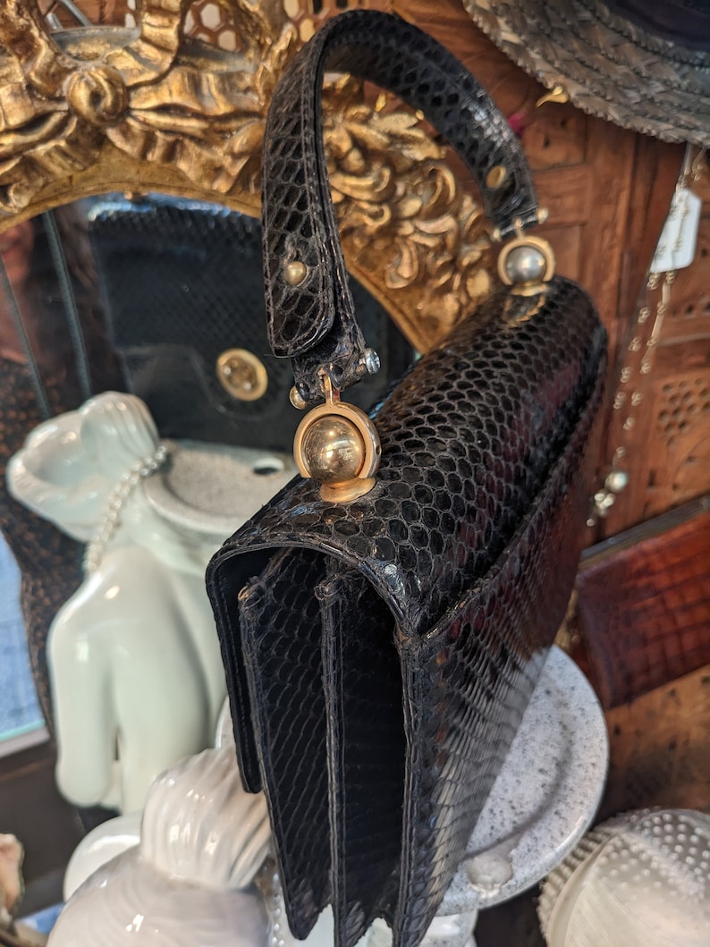 Puede incluir: Un bolso de mano de piel de serpiente negra con asa superior y herrajes dorados. El bolso tiene forma rectangular y cierre de solapa.