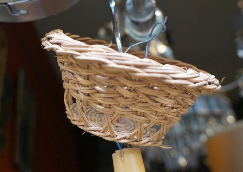 May include: A close-up of a small, woven basket made of natural materials. The basket is light brown and has a round shape. It is hanging from a wire.