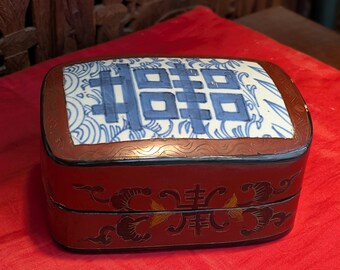 Könnte beinhalten: Eine rote und schwarze lackierte Holzkiste mit einem blauen und weißen Porzellan-Deckel. Der Deckel zeigt ein traditionelles chinesisches Schriftzeichen für "Doppeltes Glück" in Blau. Die Kiste ist mit goldenen Akzenten verziert.