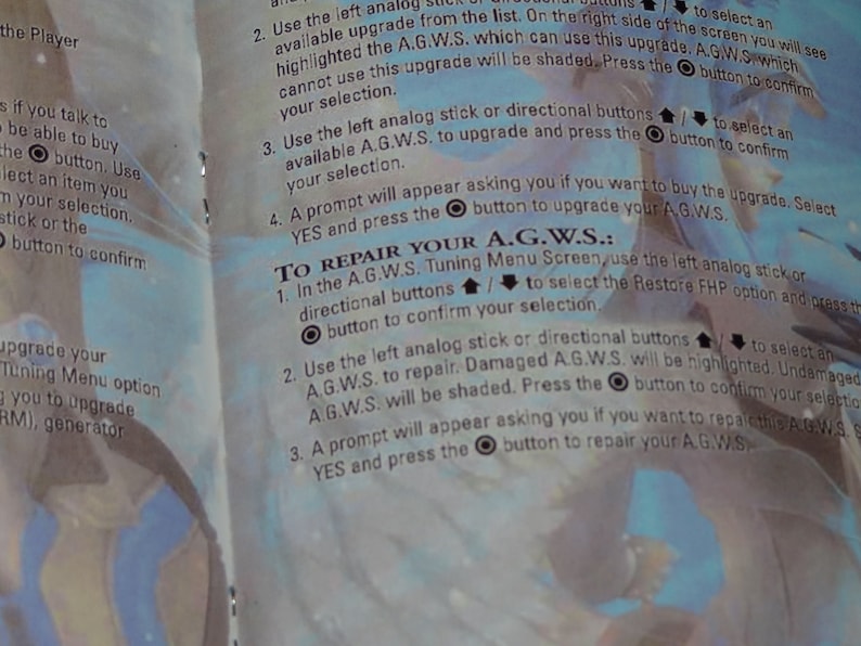 May include: A page from a video game instruction manual with a blue and white background. The text explains how to repair and upgrade the A.G.W.S. using the left analogue stick or directional buttons. The text also includes the button symbols for confirming selections.