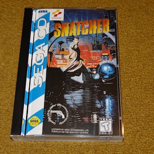 May include: A Sega CD game case with a blue and white striped spine. The front of the case features a cyberpunk-style image of a man in a trench coat holding a gun, with the title "SNATCHER" in large white letters. The game is rated "Teen" by the ESRB.
