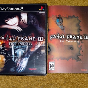 May include: A PlayStation 2 video game case and instruction manual for Fatal Frame II: The Tormented. The case features a woman with long black hair lying on a dark surface. The manual has a red and orange background with the game title in white.