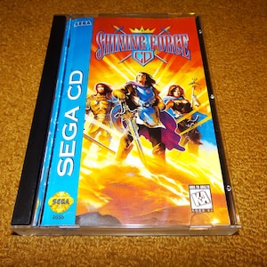 May include: A Sega CD game case with a blue background and the text "SEGA CD" in white. The game is called "Shining Force CD" and features a fantasy-themed illustration of three characters with swords and a crown.