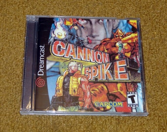 May include: A Sega Dreamcast game case with a red, yellow, and blue design. The game is called "Cannon Spike" and features a character with a yellow vest and a gun. The game is rated "Teen" by the ESRB.