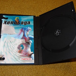 May include: A black DVD case with a white and blue cover featuring the text "Xenosaga EPISODE I Der Wille zur Macht" and a female character with blue hair and white wings. The case is open, revealing a black disc inside.