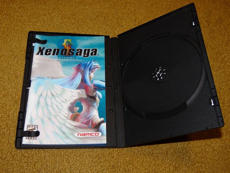 May include: A black DVD case with a white and blue cover featuring the text "Xenosaga EPISODE I Der Wille zur Macht" and a female character with blue hair and white wings. The case is open, revealing a black disc inside.